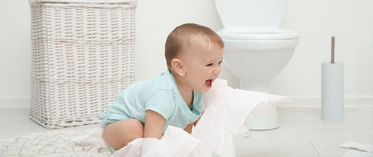 A playful baby pulls on a roll of toilet paper near a wicker basket and toilet.