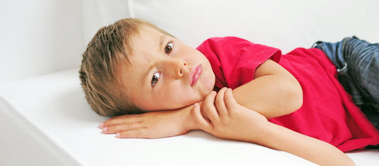  A young boy in a red shirt looks contemplative while resting his chin on his hands, lying on a white couch.