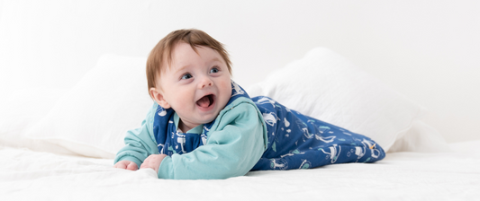 A baby dressed in pink dotted pajamas sprawls relaxed on a rumpled white bedsheet.