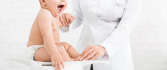 A happy baby sits on a scale during a check-up, with a healthcare professional gently holding their hand.