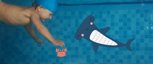 A baby wearing a blue swim cap reaches towards a cartoon crab and shark in a tiled swimming pool.