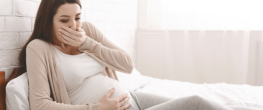  A pregnant woman sitting on a bed, covering her mouth, feeling nauseous.