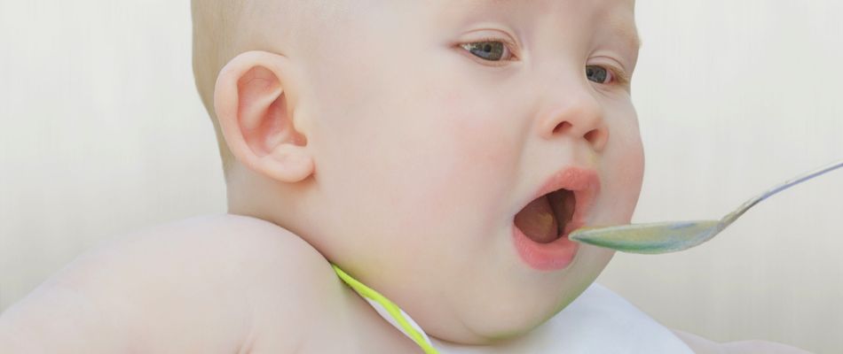  A baby opens wide for a spoonful of food, capturing an early experience of tasting solids, with a curious and eager expression.