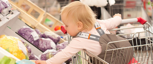 A toddler with a pacifier reaches for produce while securely strapped into a shopping cart, exploring the colorful array of vegetables at a grocery store.