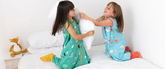  Two young girls in matching animal-themed sleep sacks playfully engage in a pillow fight on a white bed, surrounded by soft pillows and a plush fox toy.