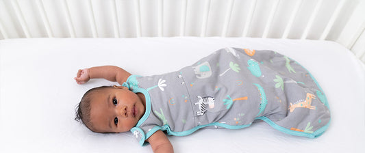 A baby lies on a white mattress, dressed in a grey sleep sack with colorful animal illustrations, looking calmly at the camera.