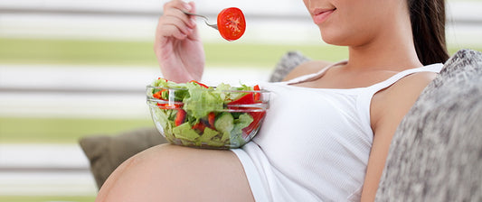 A pregnant woman reclining comfortably, eating a fresh salad with a focus on a tomato slice.