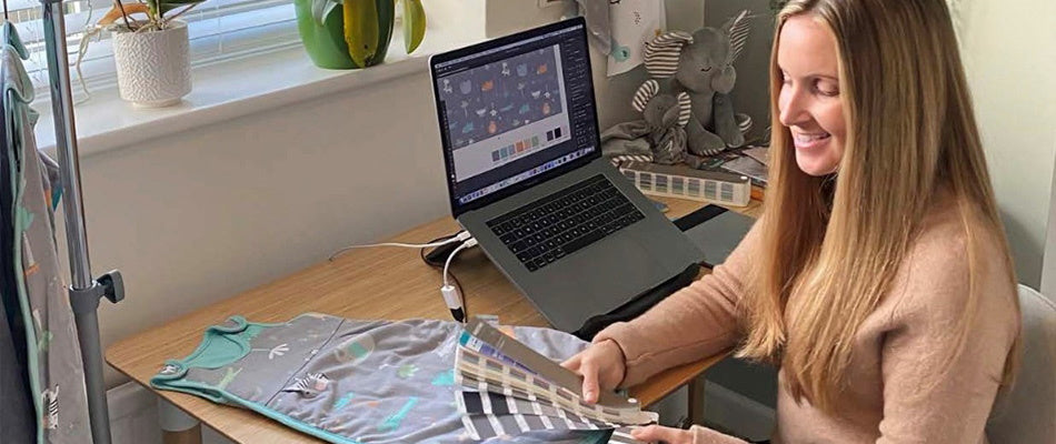 A designer smiles while working at her desk, examining fabric samples next to a laptop displaying design software, in a creatively organised workspace with children's motifs.