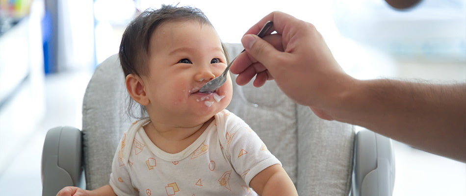 a baby in a high chair, being fed by an adult using a spoon. The baby's face is smeared with food.