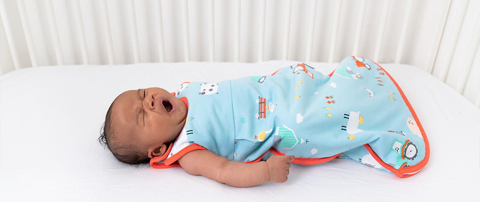 A yawning baby dressed in a colorful sleep sack lies on a white bed.