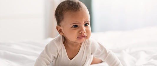  A baby with a concerned expression sits on a white bed, wearing a white onesie.