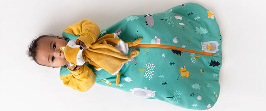 A baby dressed in a green baby sleeping bag a comforter holds toy.